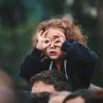 A small girls sits atop her father's sholders with her fingers circling her eyes as she pretends to see better through glasses.