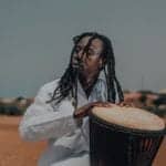 A drummer with dreadlocks, dressed in a white shirt and playing a djembe in the open air